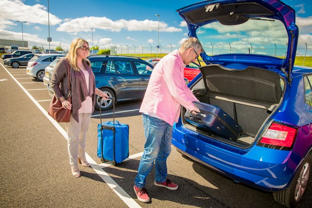 Muškarac stavlja kofer u prtljažnik plavog automobila dok žena drži ručnu torbu i kofer